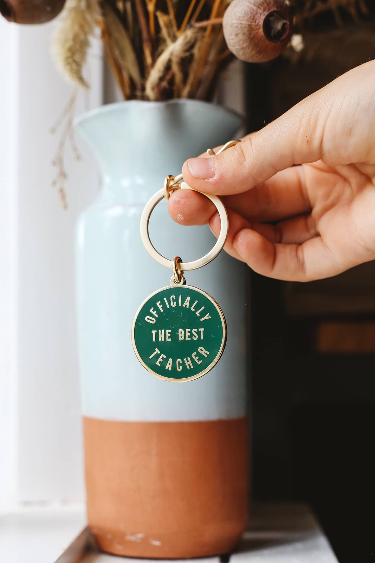 Àine & Co. Alphabet Bags metal keyring featuring circle shape with gold hardware and gold text "Officially The Best Teacher" on dark green background