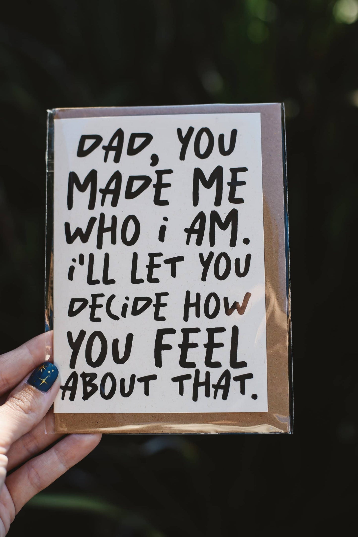 Card with text "Dad, you made me who I am. I'll let you decide how you feel about that" held by a hand against a dark background