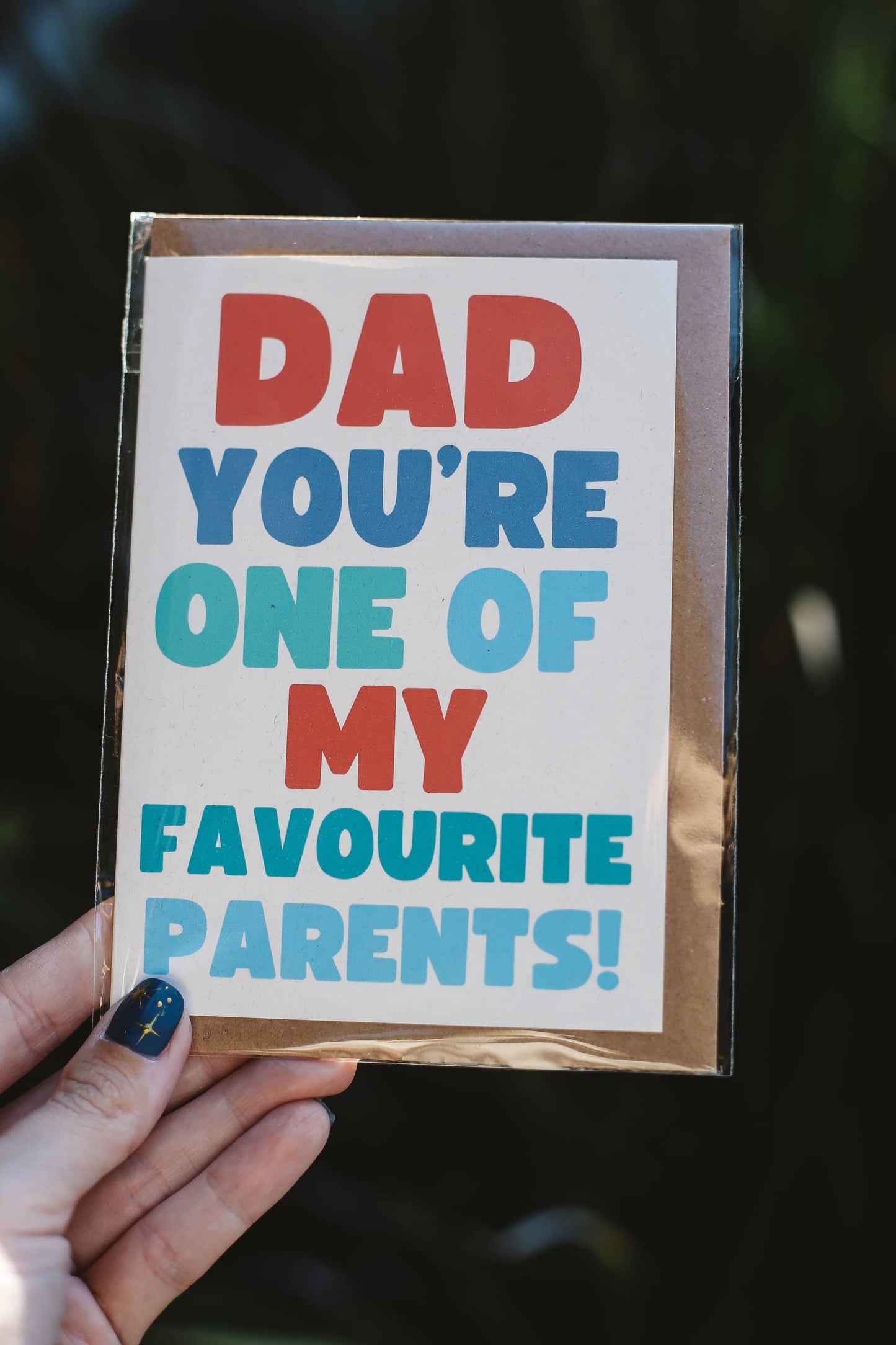 Card with colorful text 'Dad you're one of my favourite parents!' held by a hand against a dark background.