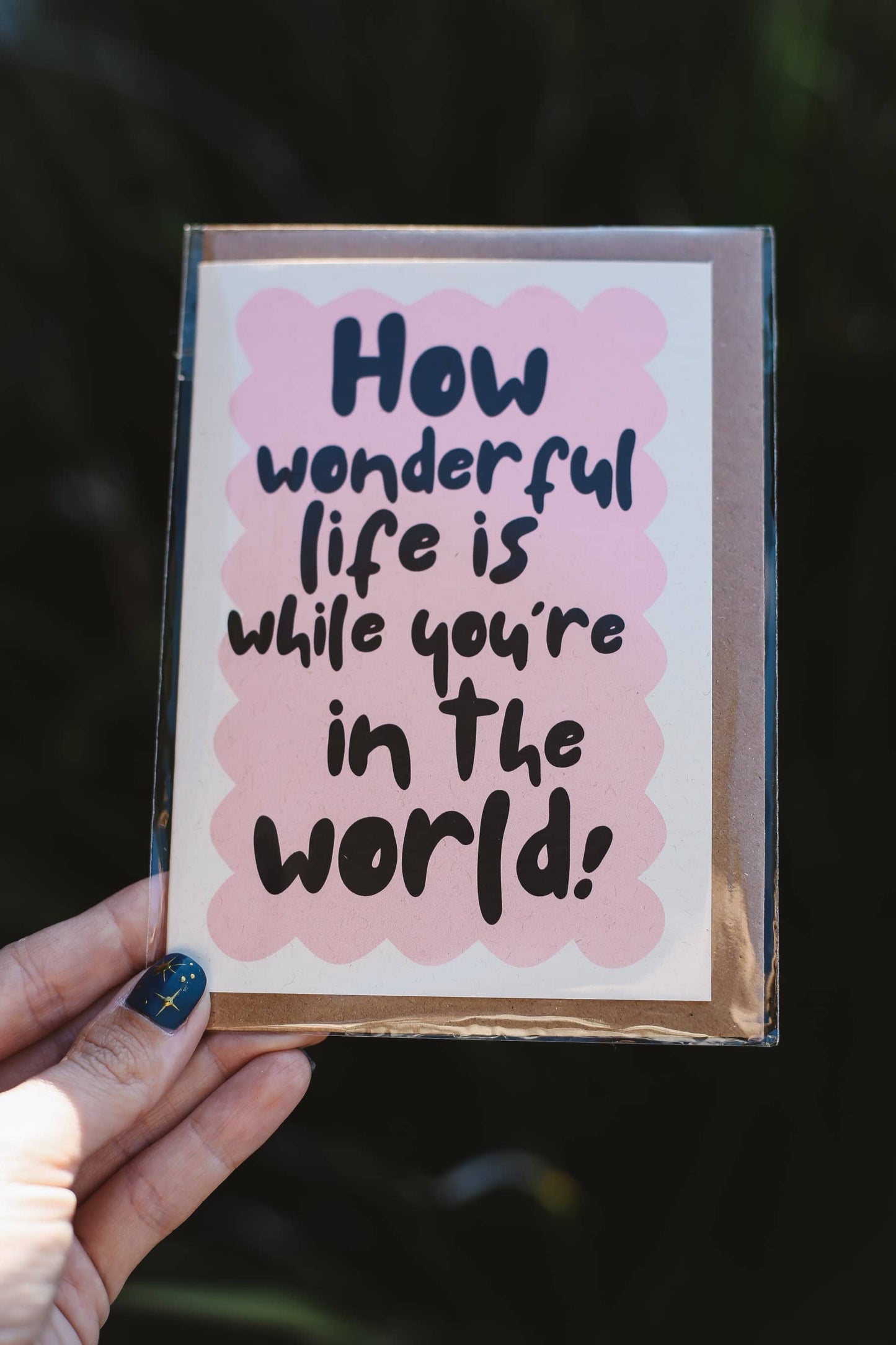 Hand holding a card with 'How wonderful life is while you're in the world!' text on a pink background.