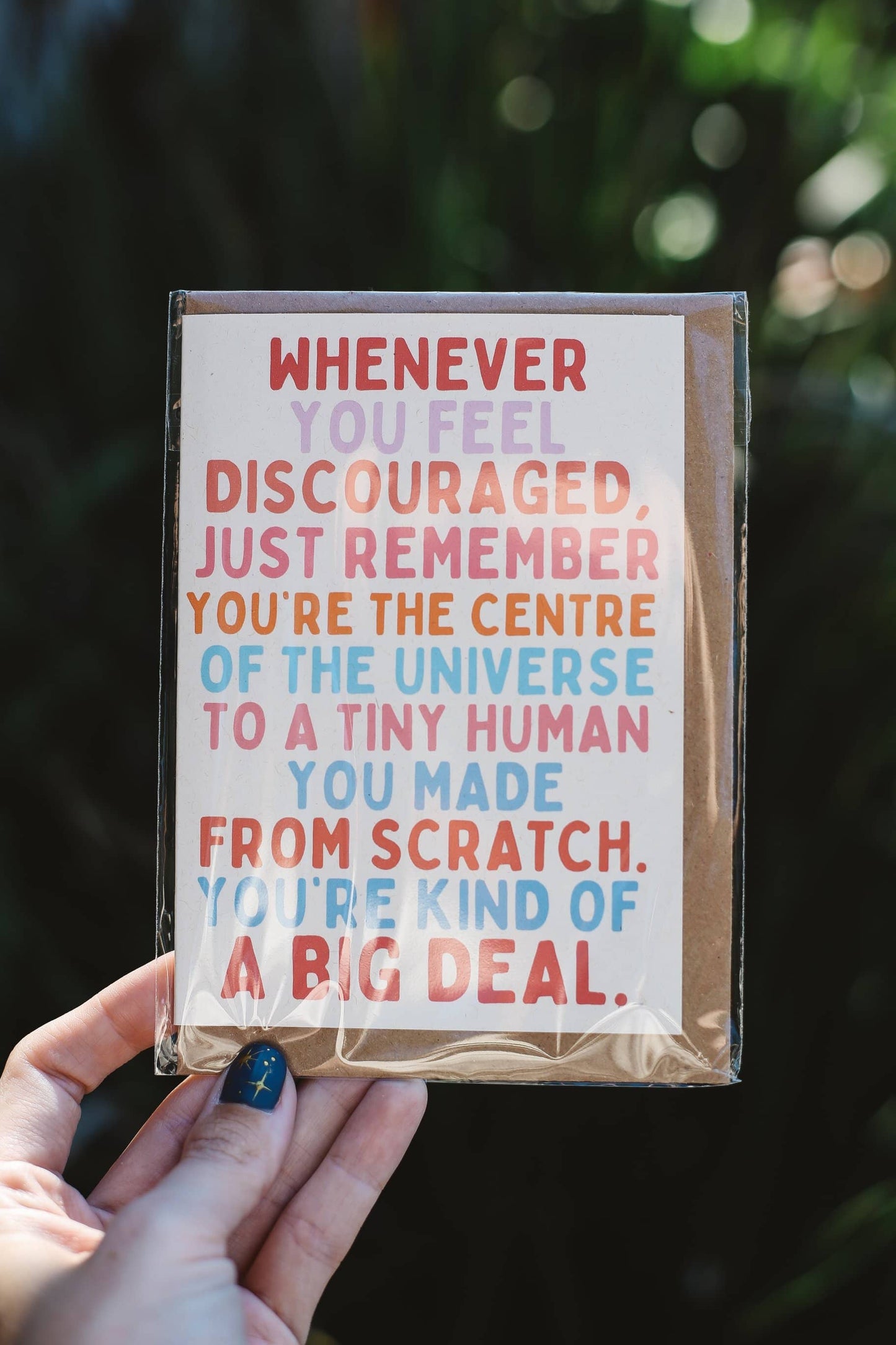 Card with text "Whenever you feel discouraged, just remember you're the centre of the universe to a tiny human you made from scratch. You're kind of a big deal" held by a hand against a blurred natural background
