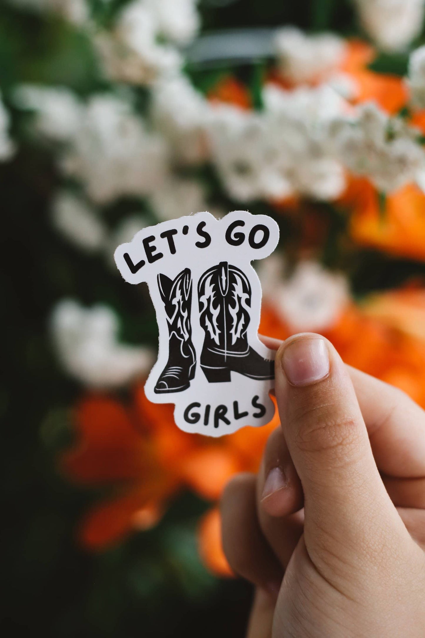Hand holding a sticker with cowboy boots and 'Let's Go Girls' text against a floral background
