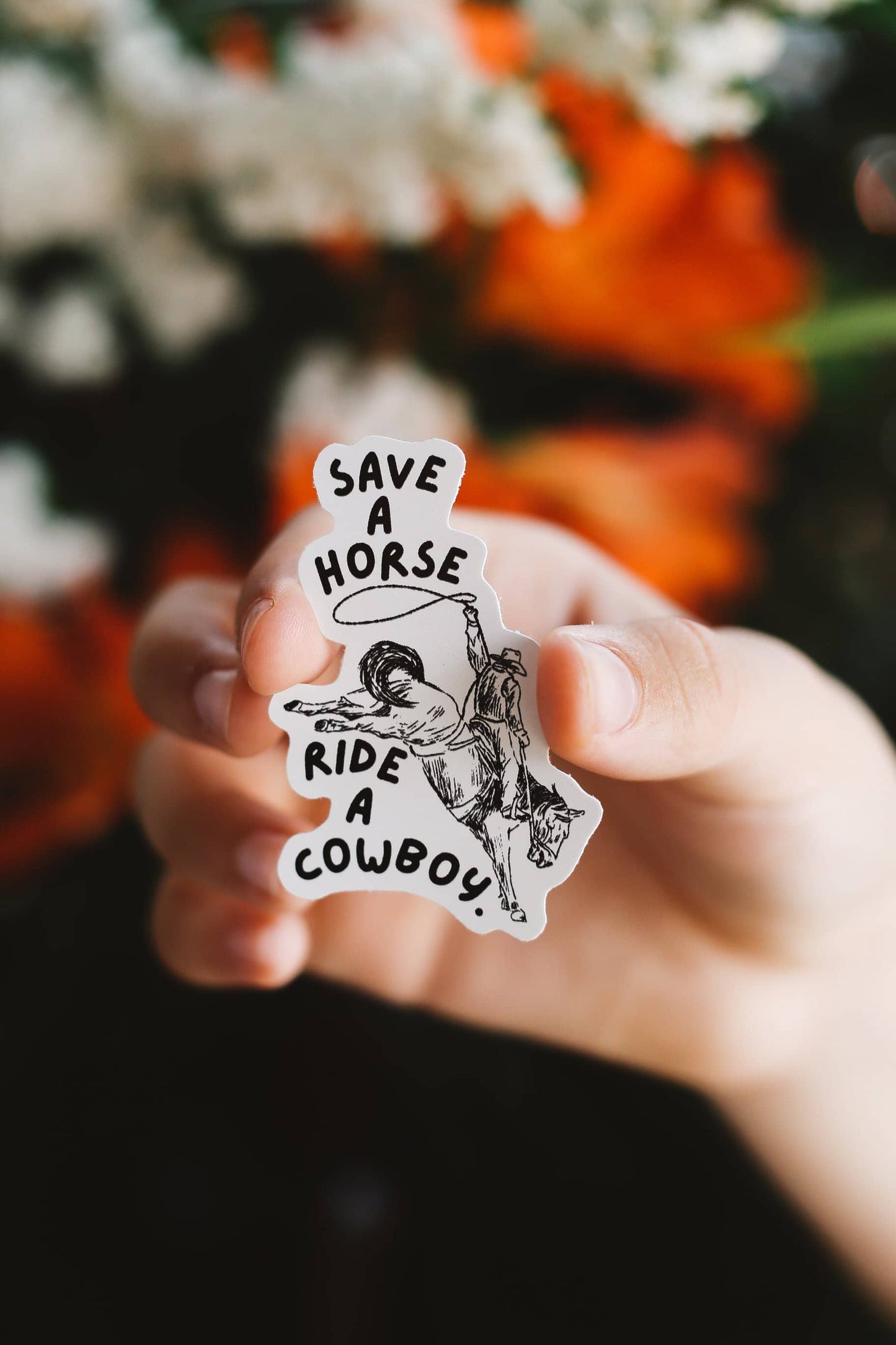 Hand holding a sticker with text 'Save a horse, ride a cowboy' against a blurred floral background