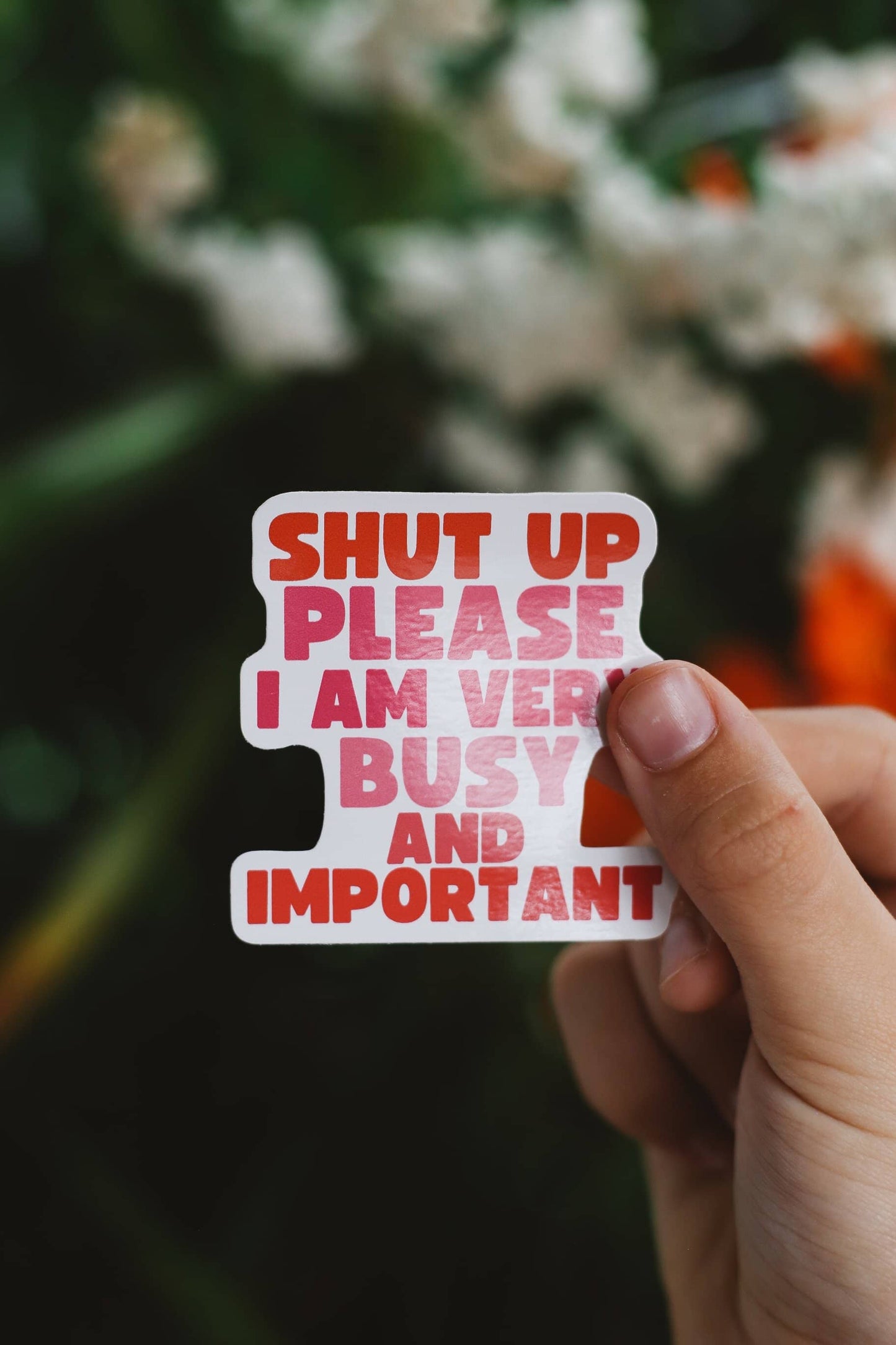 Hand holding a sticker with text 'SHUT UP PLEASE I AM VERY BUSY AND IMPORTANT' against a blurred natural background.