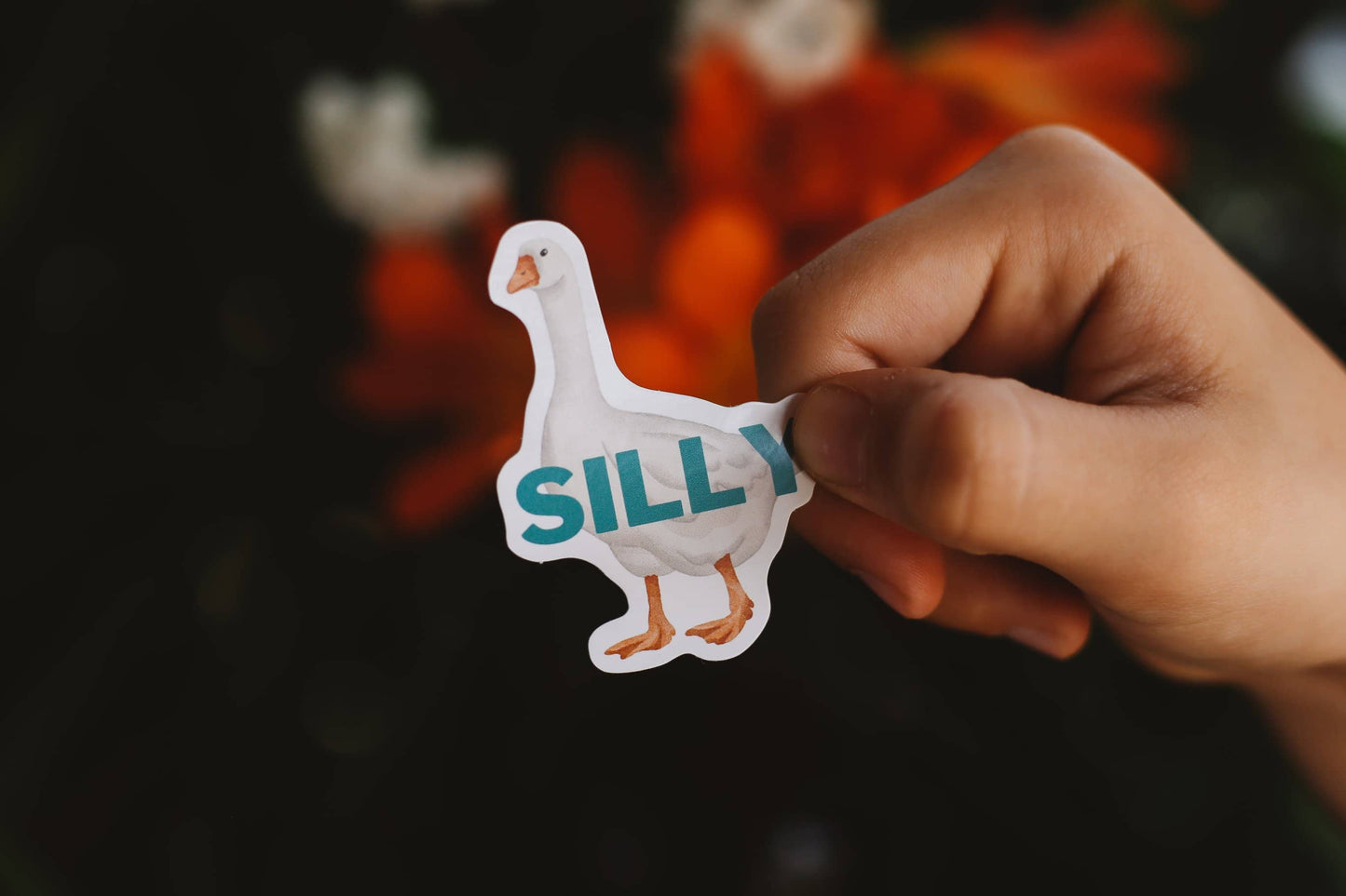 Hand holding a sticker with a goose design and 'SILLY' text against a blurred background