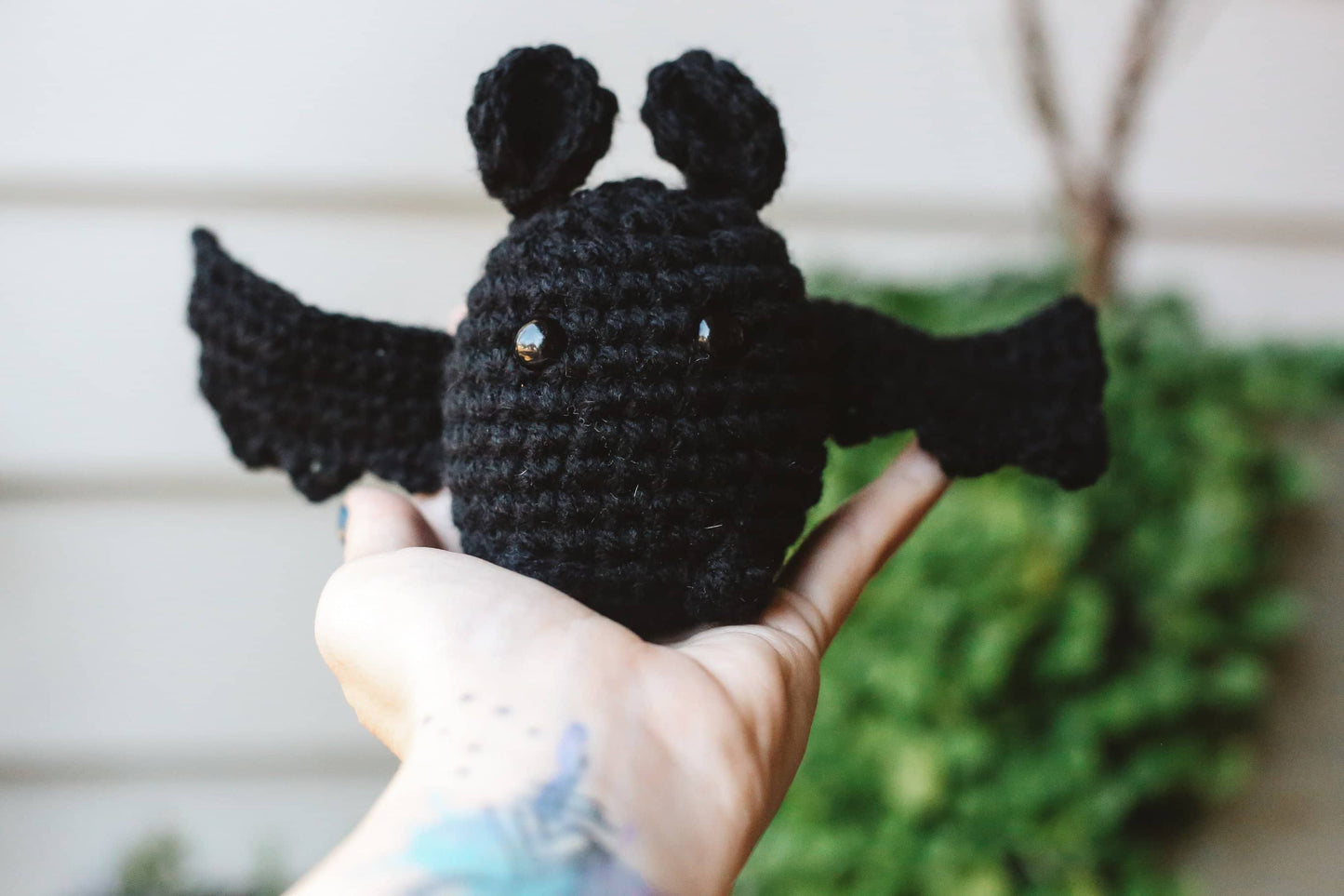 Black crocheted bat toy held in a hand with a blurred green plant background