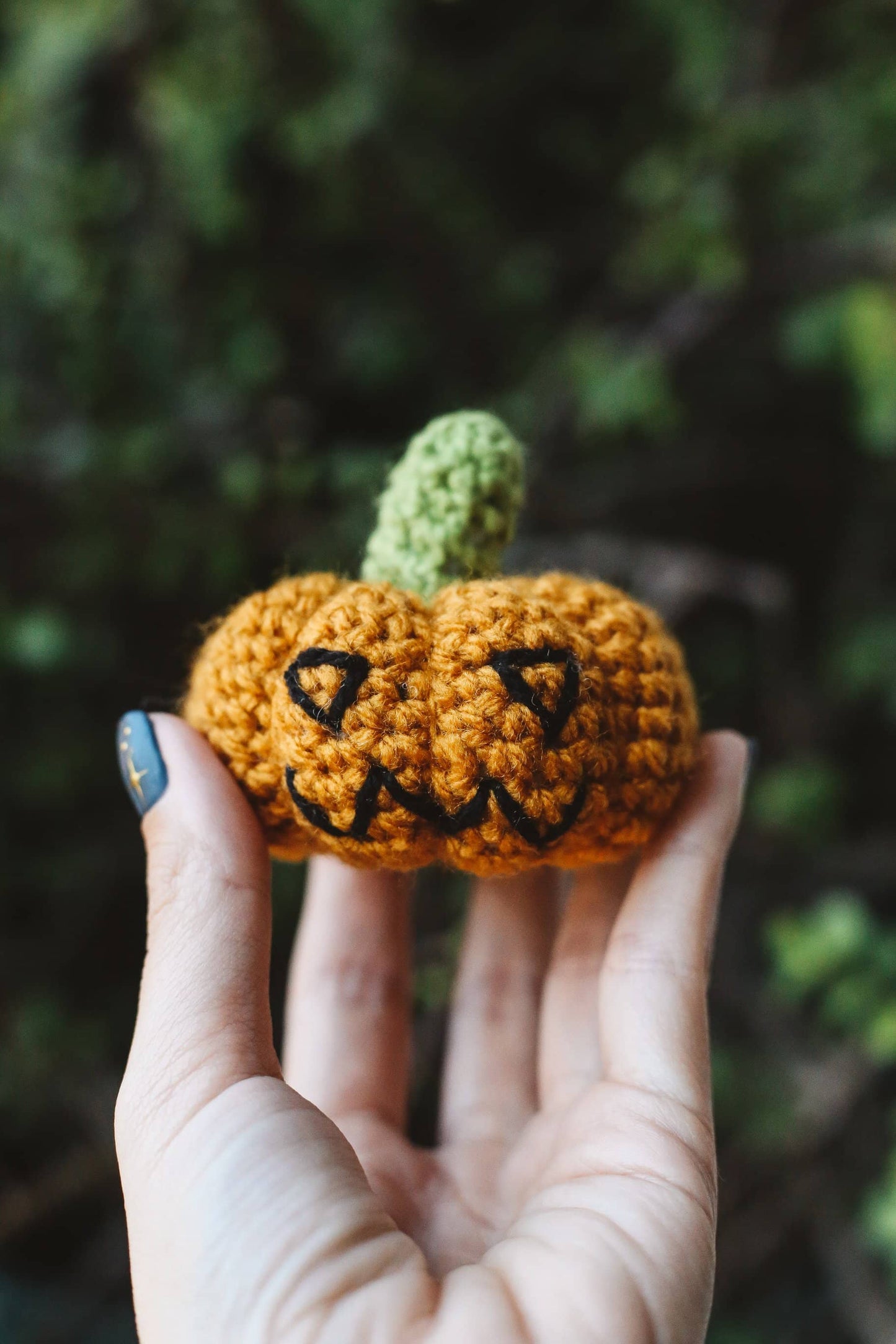 Hand holding a small crocheted pumpkin with a face against a blurred natural background
