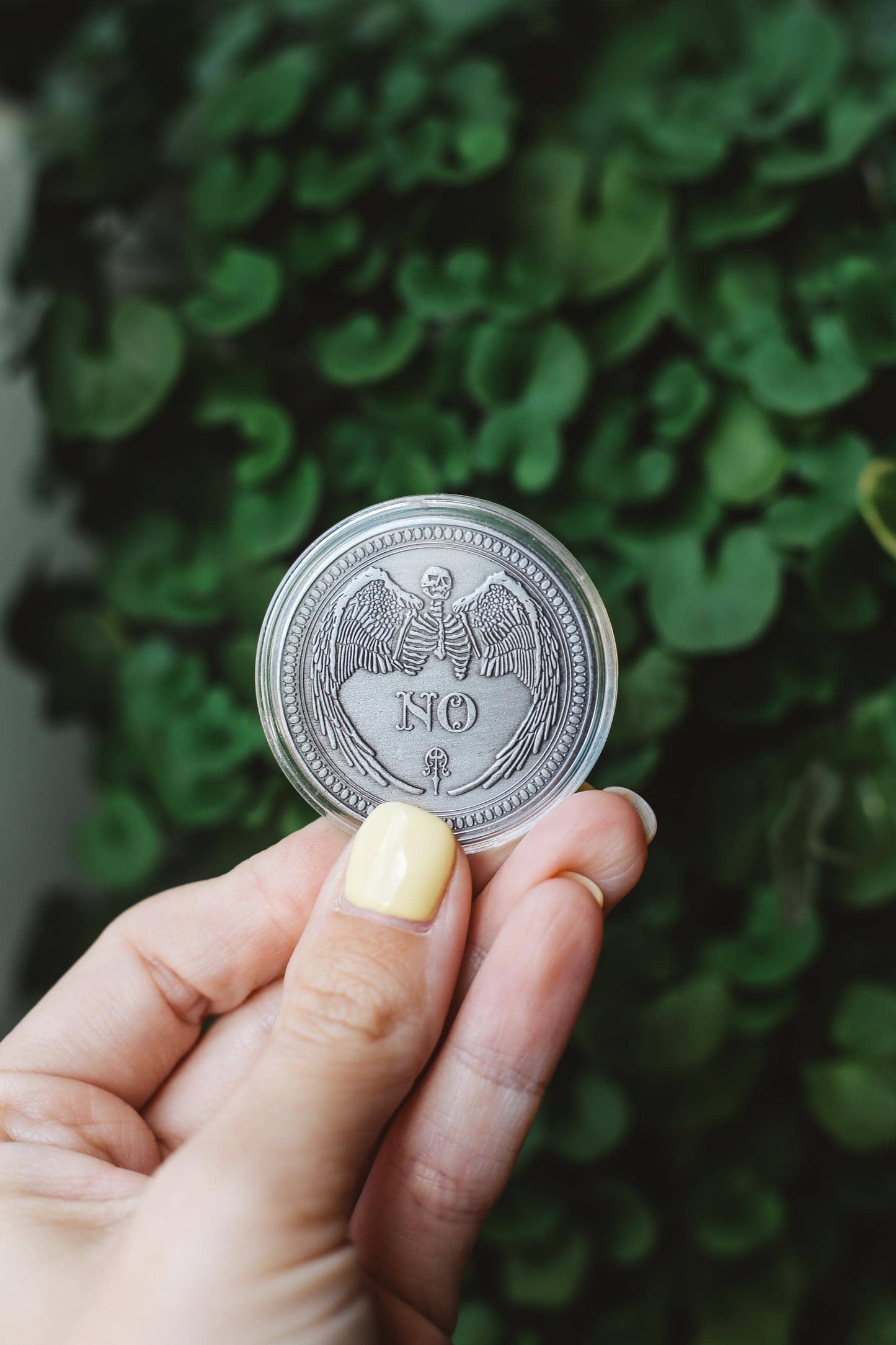 Hand holding a silver coin with 'NO' engraving featuring winged skeleton against a green leafy background