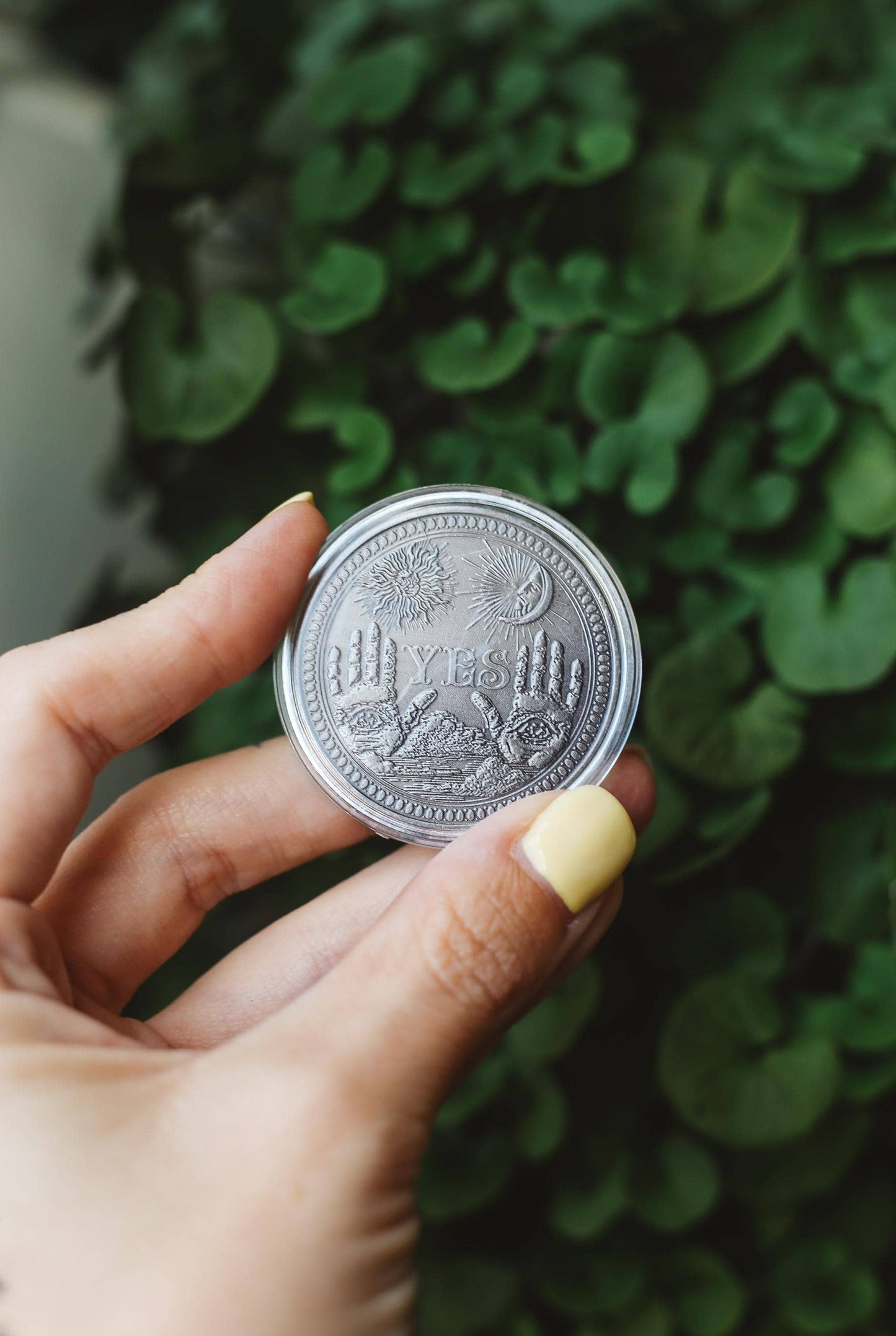 Hand holding a silver coin with embossed mystical design against a green leafy background