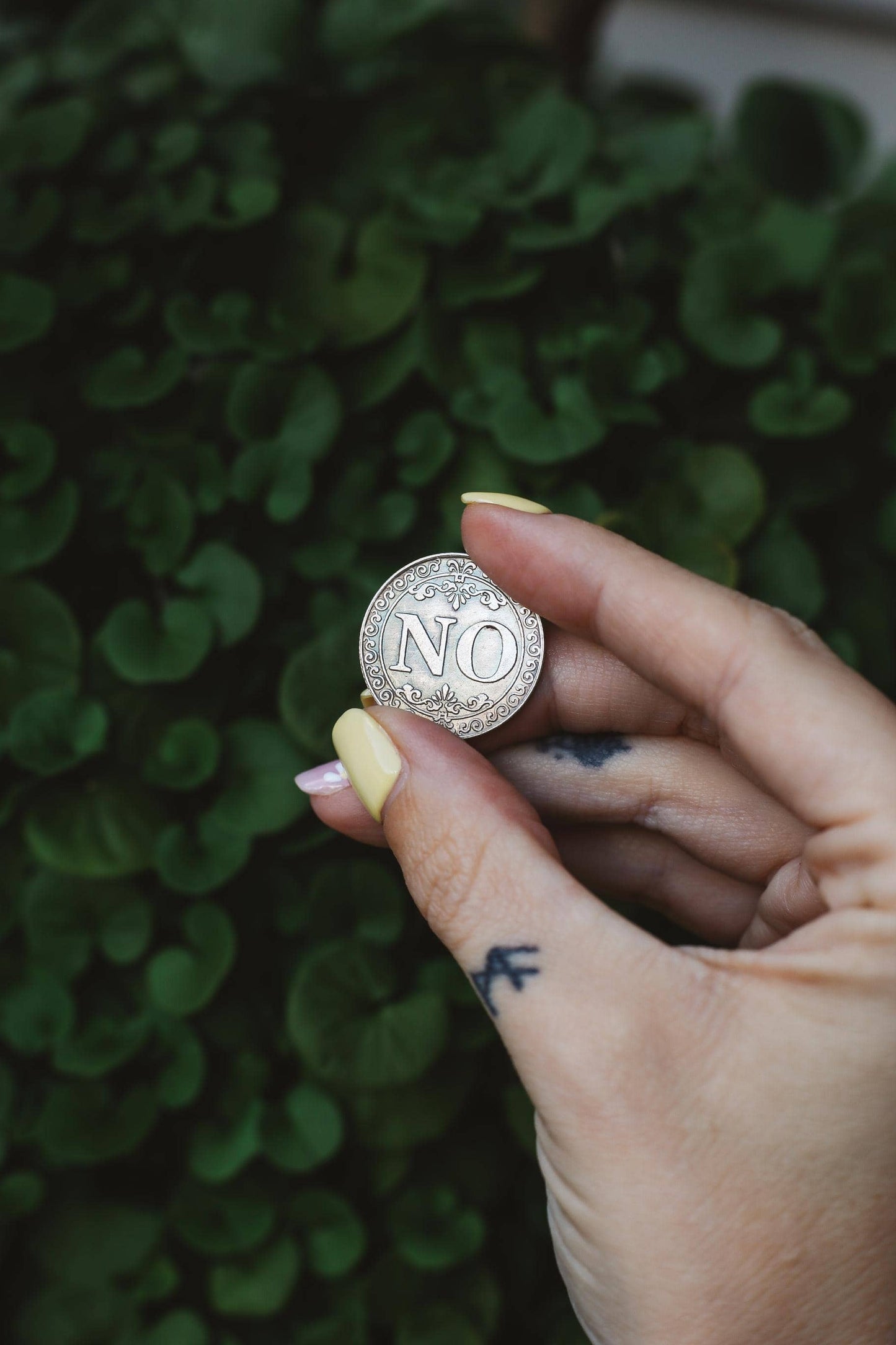 Hand holding a silver coin with 'NO' engraving against a green leafy background