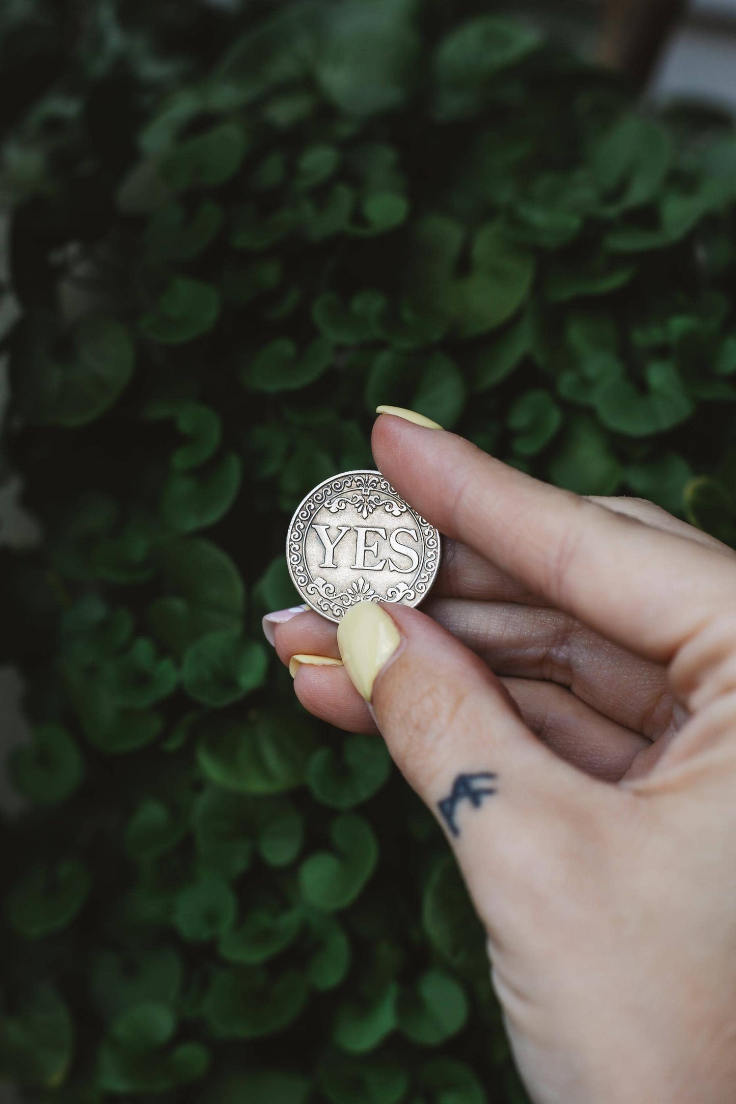 Hand holding a silver coin with 'YES' on it against a green leafy background