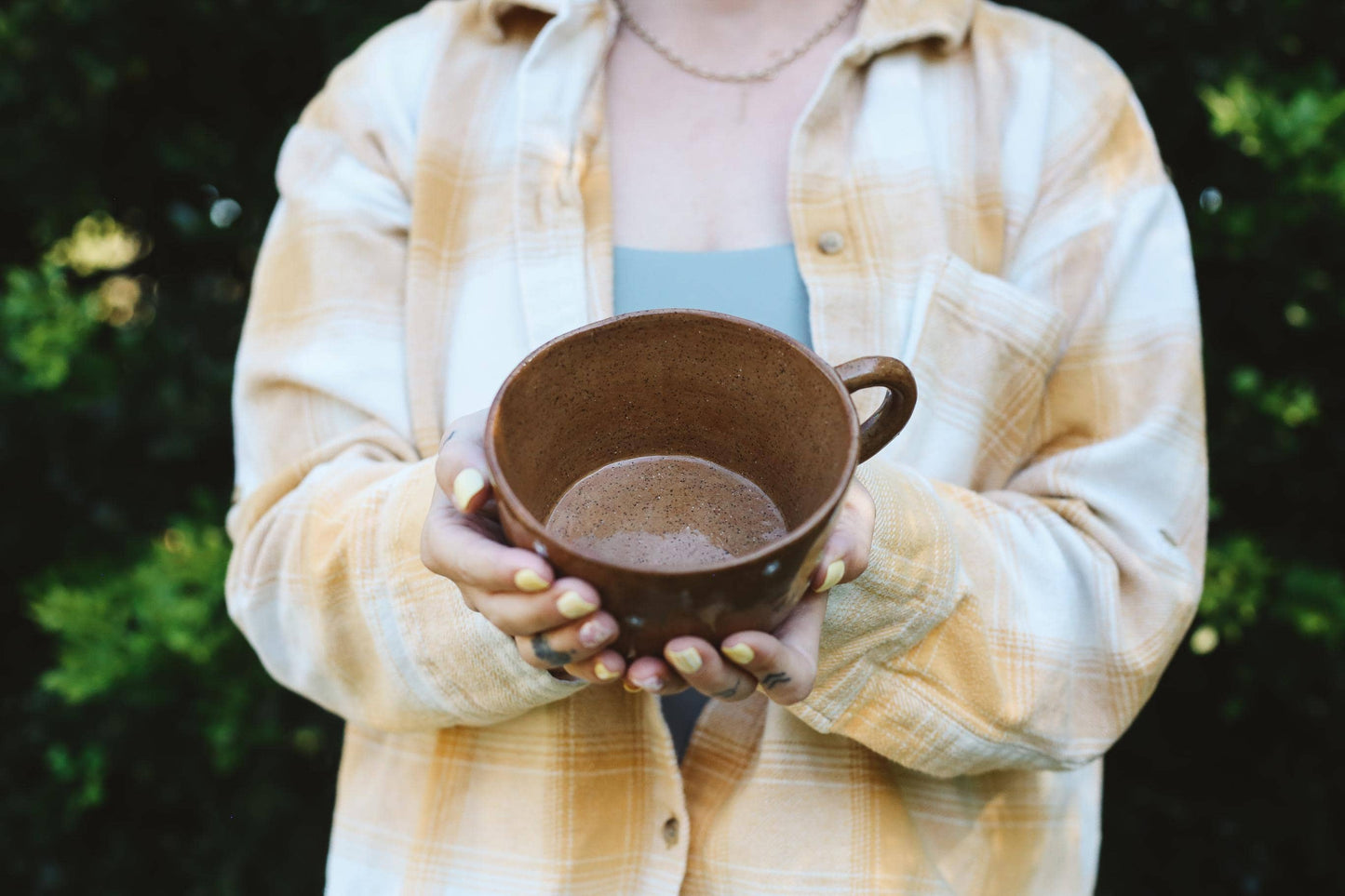 Person holding a brown ceramic cup outdoors with greenery in the background