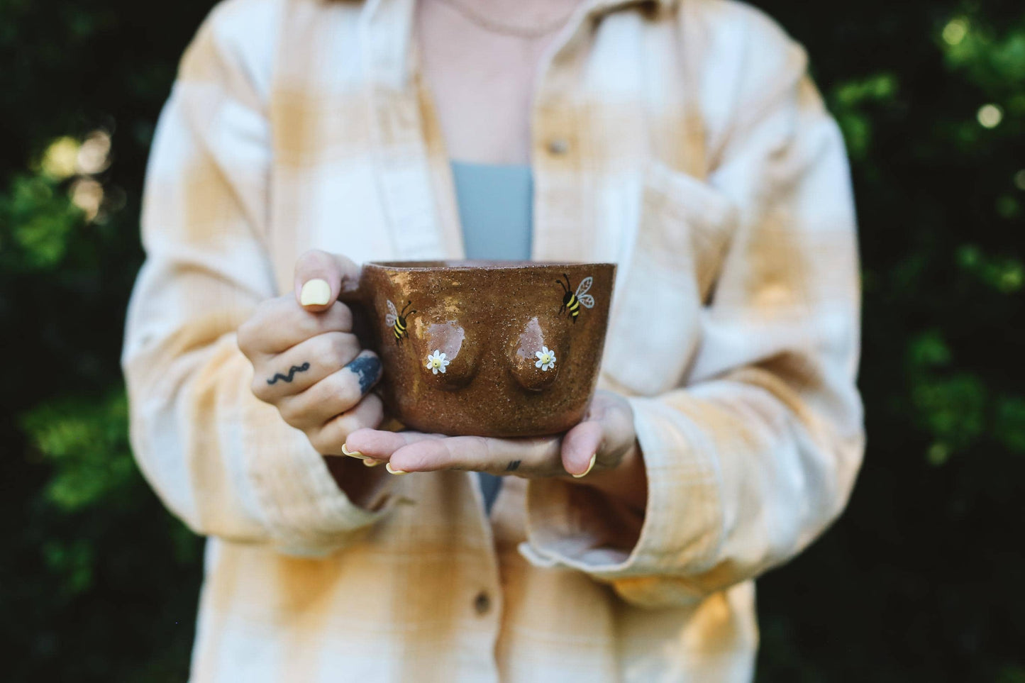 Person holding a ceramic mug with boob shapes and bee design elements against a blurred natural background