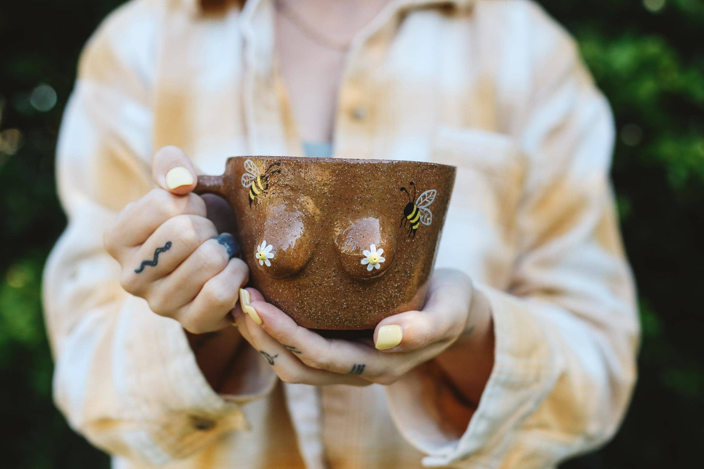 Person holding a brown mug with a boob and bumblebee design against a blurred green background
