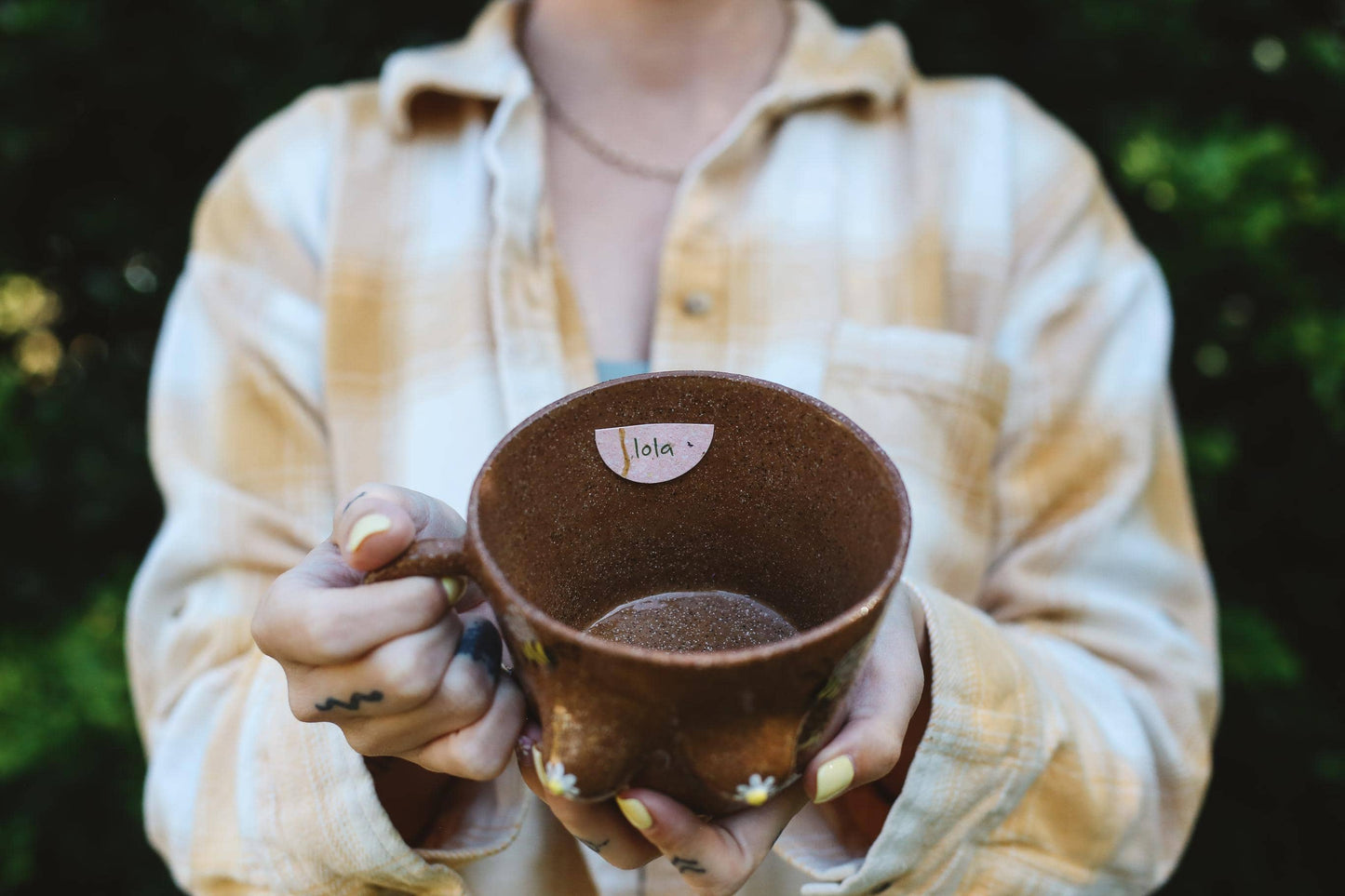 Person holding a brown ceramic mug with name tag "Lola" against a blurred natural background