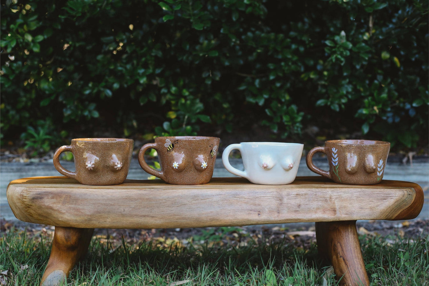 Four ceramic mugs on a wooden stool outdoors with greenery in the background