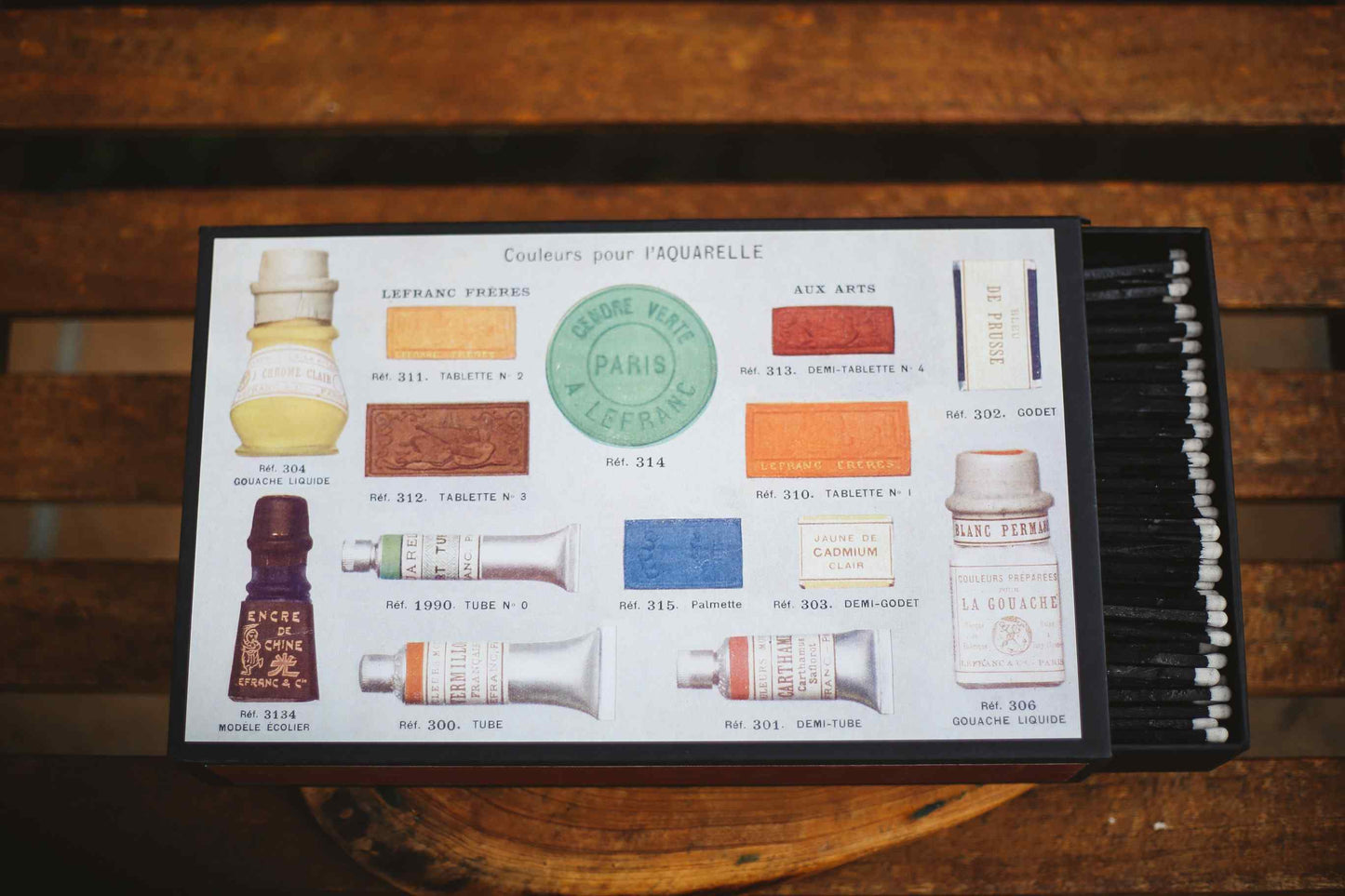 A vintage-style hair color palette card from Paris Esthetique, displayed on a rustic wooden surface. The card features illustrations of various hair dye bottles, color swatches, and a row of black applicator brushes on the right side.