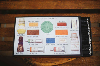 A vintage-style hair color palette card from Paris Esthetique, displayed on a rustic wooden surface. The card features illustrations of various hair dye bottles, color swatches, and a row of black applicator brushes on the right side.