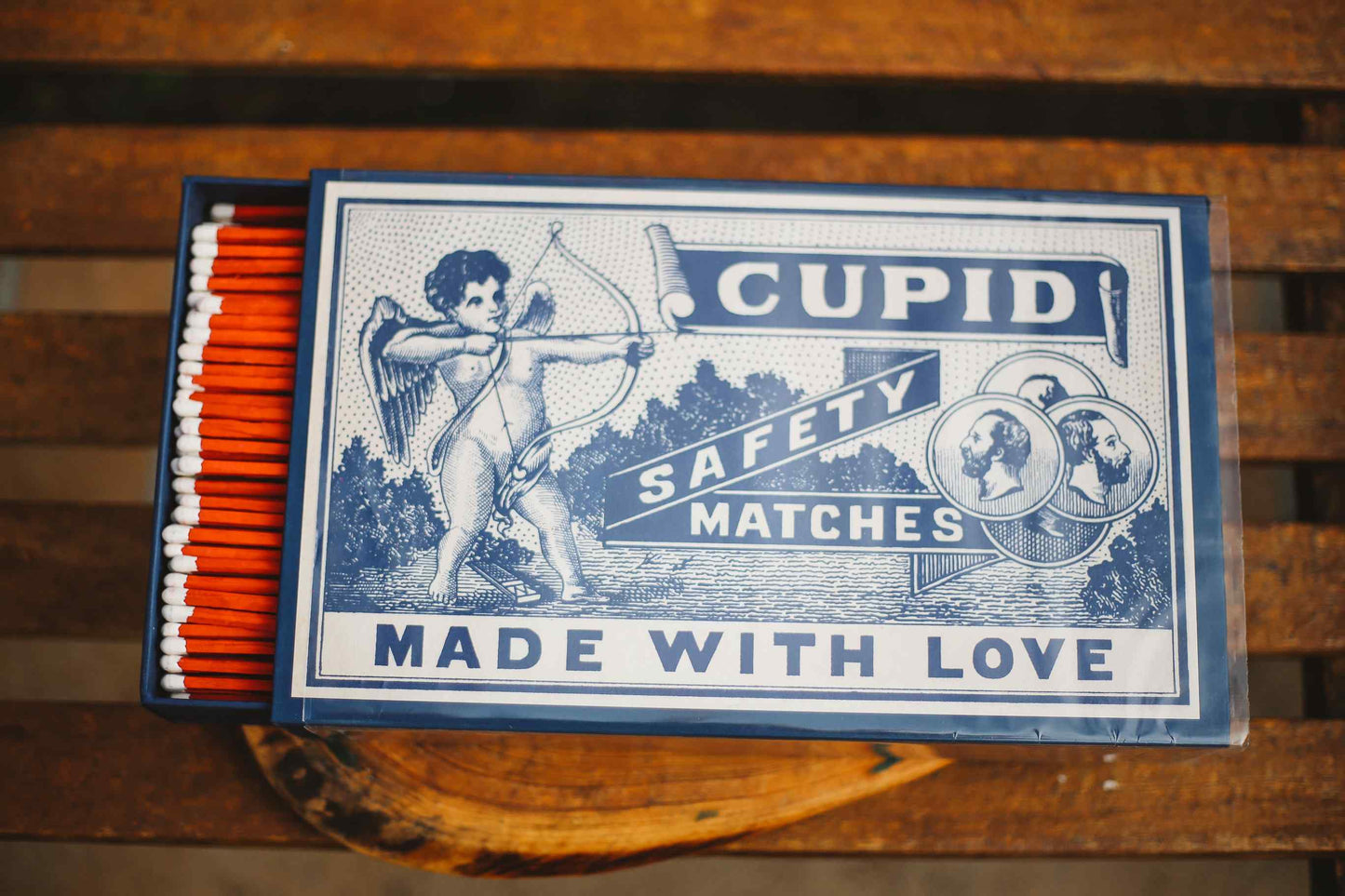 A vintage-style matchbox labeled 'CUPID SAFETY MATCHES MADE WITH LOVE' in blue and white, with orange-red matches visible in the drawer, placed on a wooden surface.