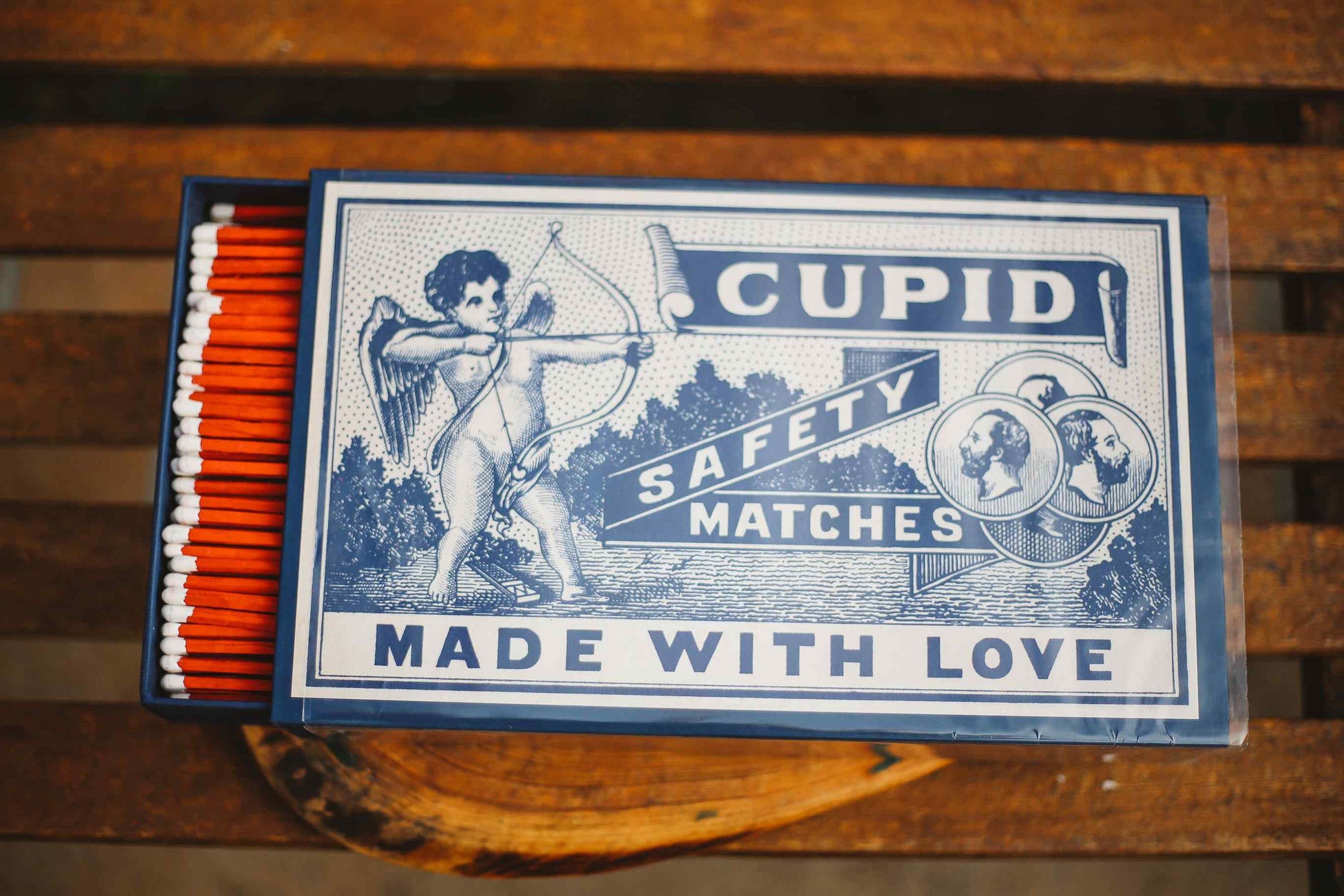 A vintage-style matchbox labeled 'CUPID SAFETY MATCHES MADE WITH LOVE' in blue and white, with orange-red matches visible in the drawer, placed on a wooden surface.