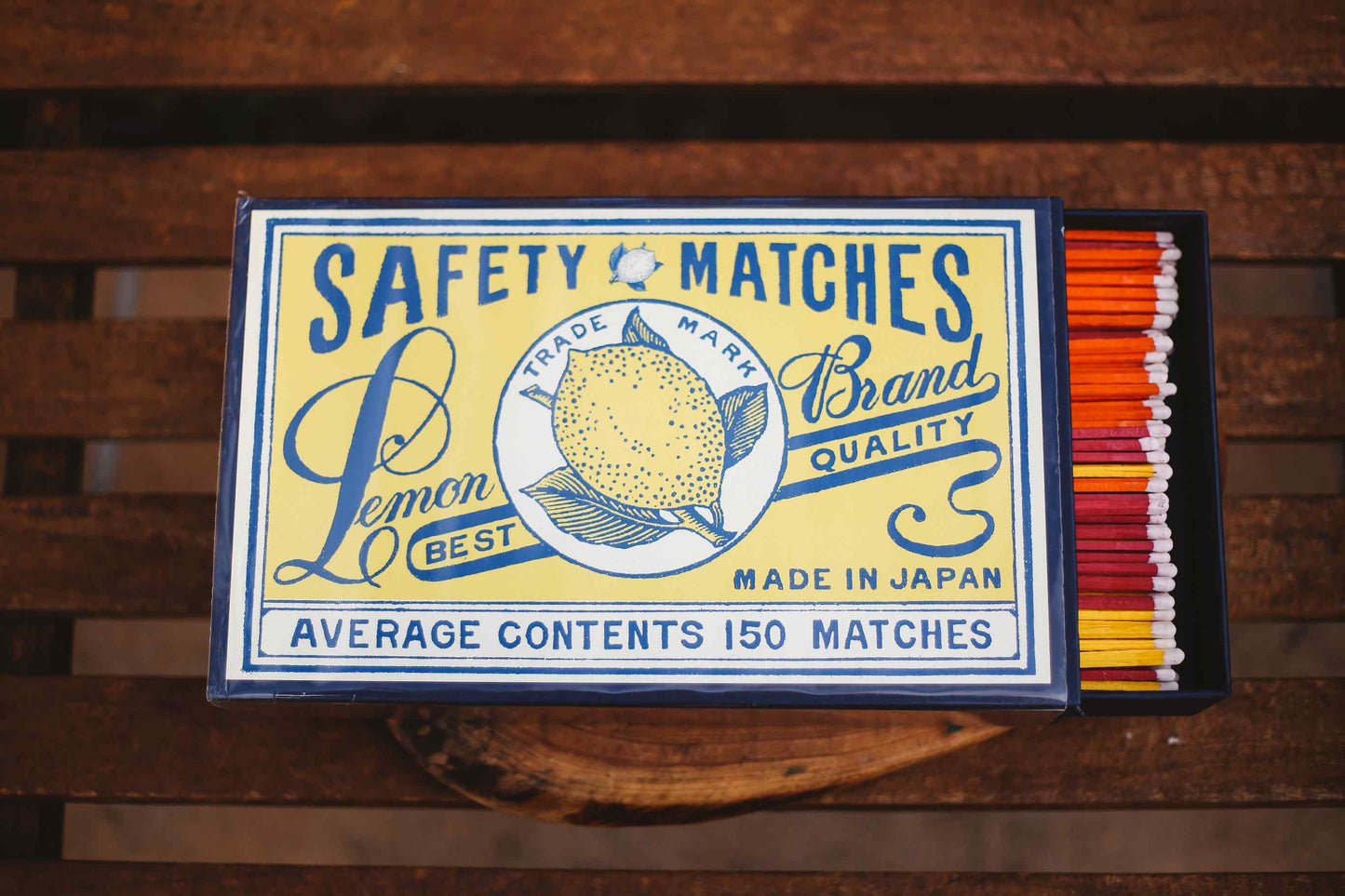 A vintage-style matchbox labeled 'SAFETY MATCHES' with a lemon illustration and text including 'Lemon Brand Quality' and 'Made in Japan,' shown open on a wooden surface with orange, red, yellow, and white matches visible inside.
