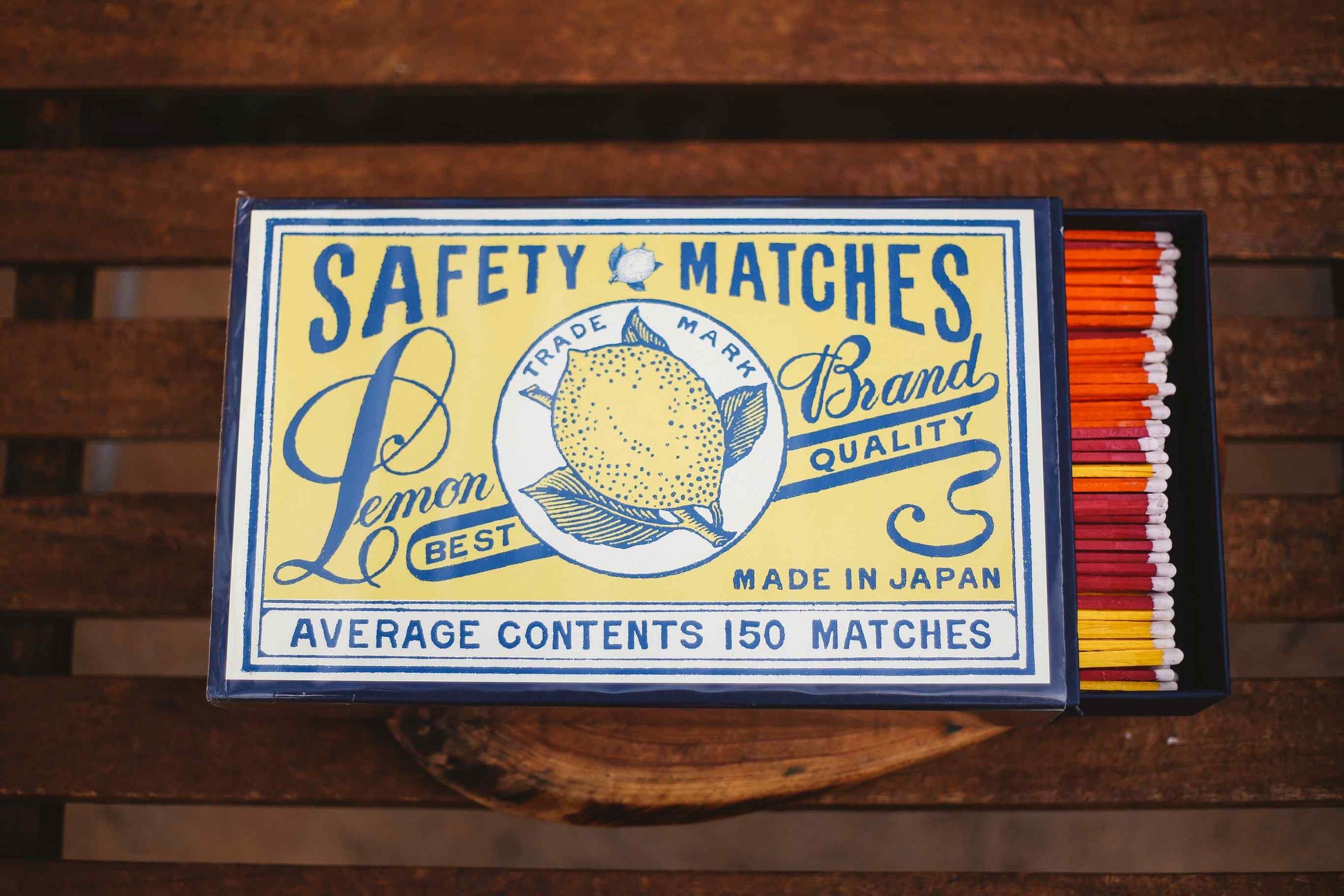 A vintage-style matchbox labeled 'SAFETY MATCHES' with a lemon illustration and text including 'Lemon Brand Quality' and 'Made in Japan,' shown open on a wooden surface with orange, red, yellow, and white matches visible inside.