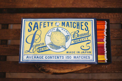 A vintage-style matchbox labeled 'SAFETY MATCHES' with a lemon illustration and text including 'Lemon Brand Quality' and 'Made in Japan,' shown open on a wooden surface with orange, red, yellow, and white matches visible inside.