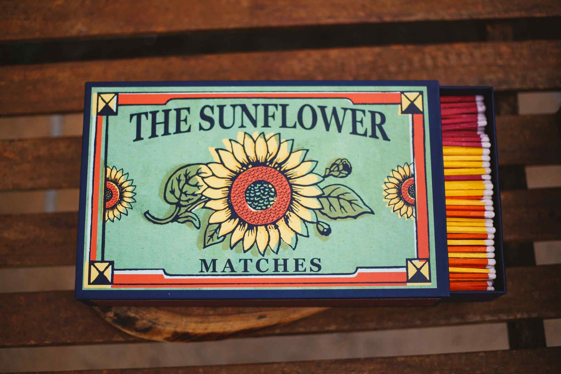 A decorative matchbox labeled 'THE SUNFLOWER MATCHES' sits on a wooden surface. The front cover features a large sunflower illustration on a light blue-green background with red, yellow, and navy accents.