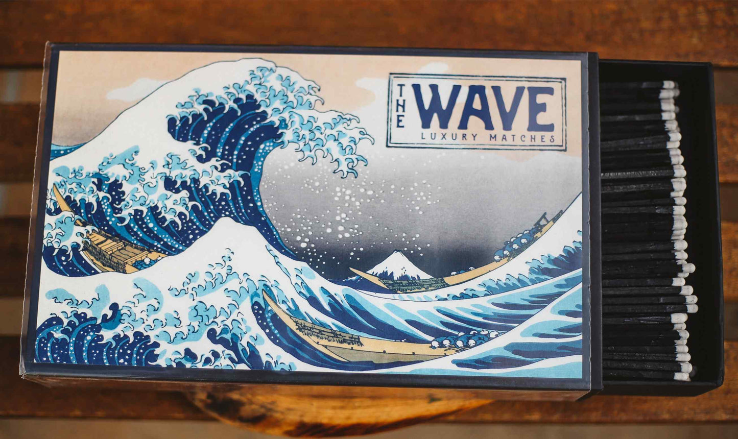 A matchbox-style product featuring a printed illustration of 'The Wave' from Katsuichi Ogata's Great Wave off Kanagawa. The left side shows the illustrated front with the text 'THE WAVE LUXURY MATCHES,' and the right side reveals a row of black matches with white tips inside a dark box, set on a wooden surface.