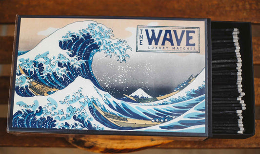 A matchbox-style product featuring a printed illustration of 'The Wave' from Katsuichi Ogata's Great Wave off Kanagawa. The left side shows the illustrated front with the text 'THE WAVE LUXURY MATCHES,' and the right side reveals a row of black matches with white tips inside a dark box, set on a wooden surface.