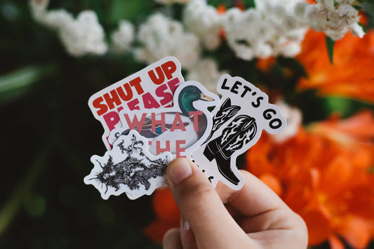 Hand holding three stickers with text and graphics against a floral background