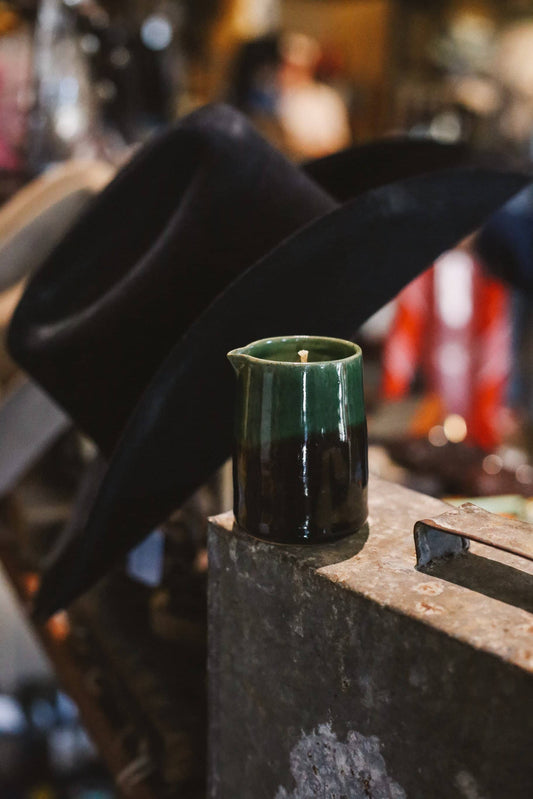 Green ceramic mug with a candle on a metal surface, with a cowboy hat in the background.