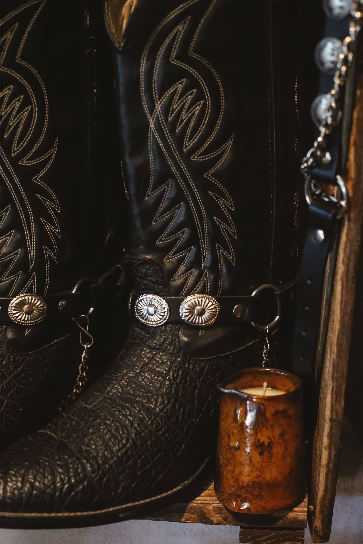 Close-up of black cowboy boots with intricate designs, next to a candle in a ceramic milk jug.