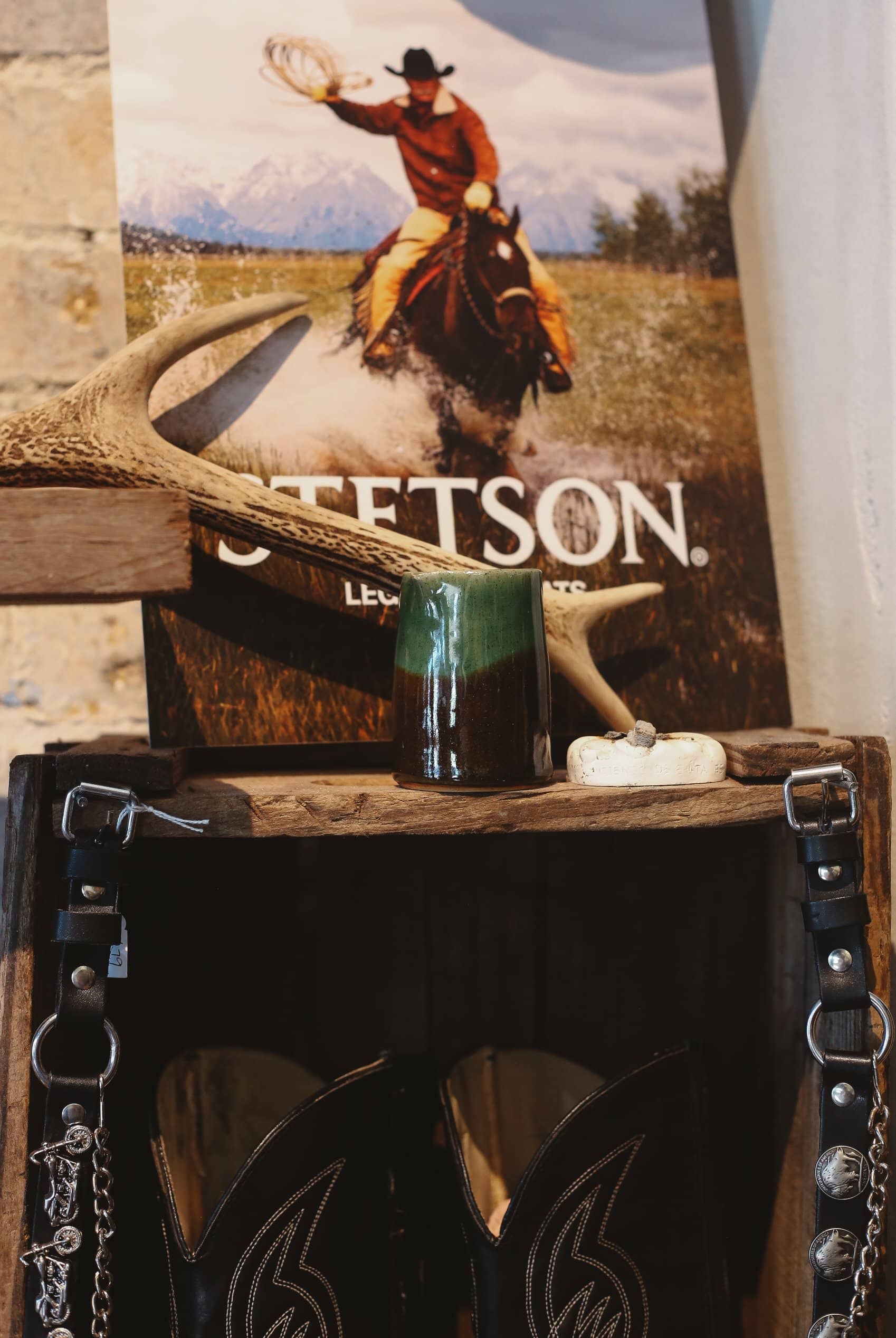 Decorative display with a body balm candle, antlers, and a Stetson poster on a wooden surface with cowboy boots underneath
