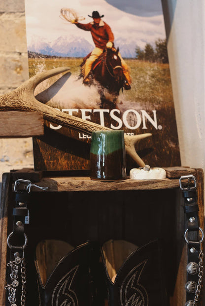 Decorative display with a body balm candle, antlers, and a Stetson poster on a wooden surface with cowboy boots underneath