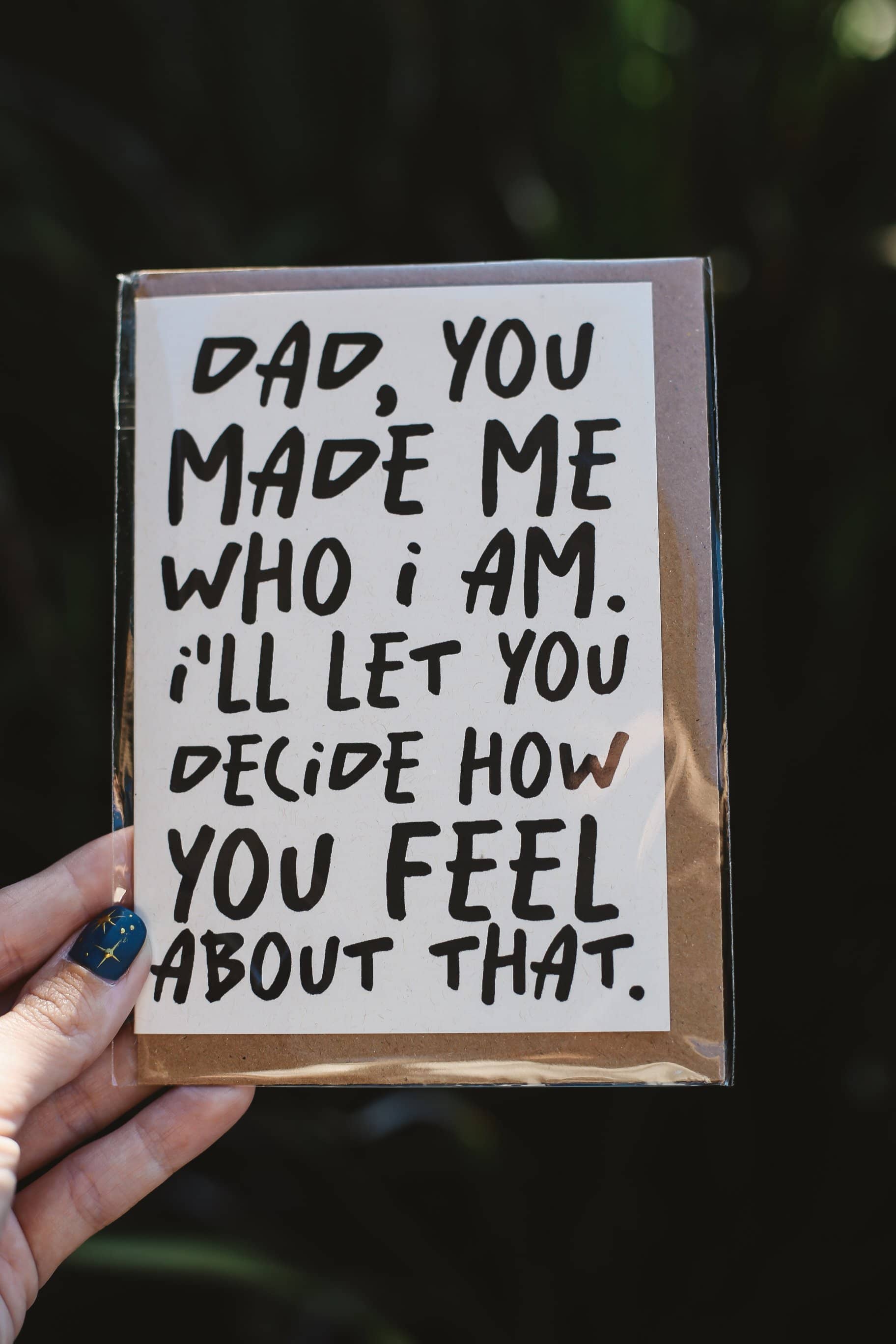 Card with text "Dad, you made me who I am. I'll let you decide how you feel about that" held by a hand against a dark background