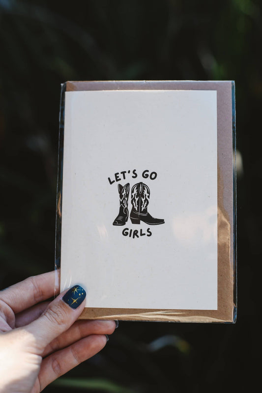 Card with 'Let's Go Girls' text and boots held by a hand against a dark background