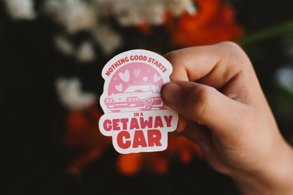 Hand holding a sticker with text 'Nothing good starts in a getaway car' against a blurred background