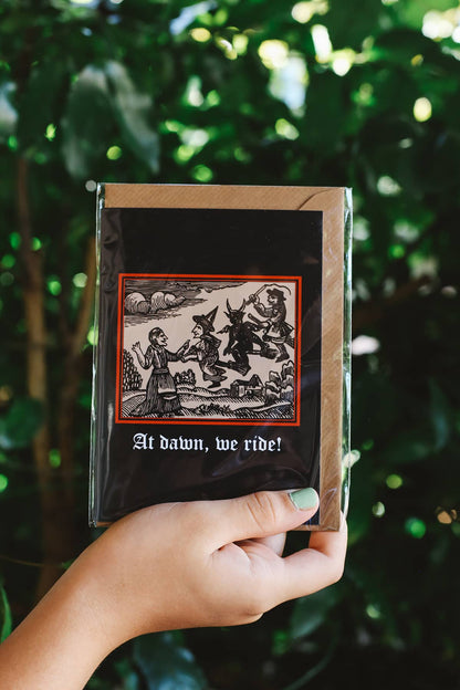 Clavis & Claustra greeting card featuring black background and witch illustration with graphic text "At dawn, we ride!"