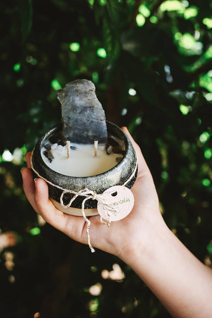 Hand holding Àine & Co. Crystal Comfort Candle in Lupercalia, featuring ceramic black and grey glazed mug, with large grey crystal atop the candle wax