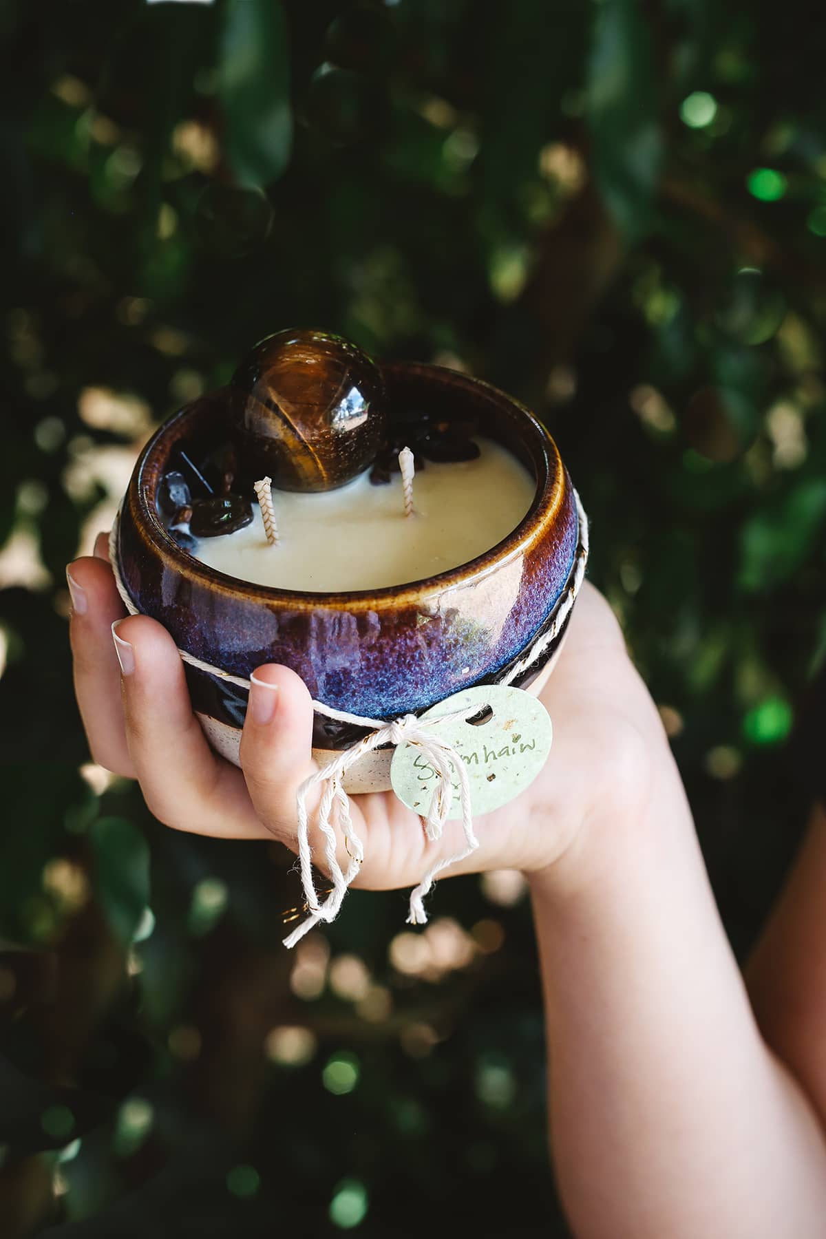 Hand holding Àine & Co. Crystal Comfort Candle in Samhain, featuring ceramic blue, purple and gold glazed mug, with tiger eye crystal atop the candle wax
