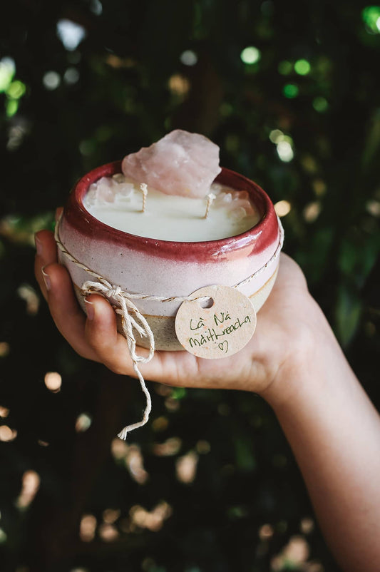 Hand holding Àine & Co. Crystal Comfort Candle in La Na Maithreacha, featuring ceramic red and pink glazed mug, with rose quartz atop the candle wax