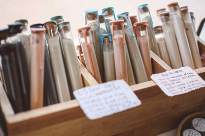 A wooden display box filled with many small glass tubes or vials containing different colored substances, with handwritten notes attached to the front.