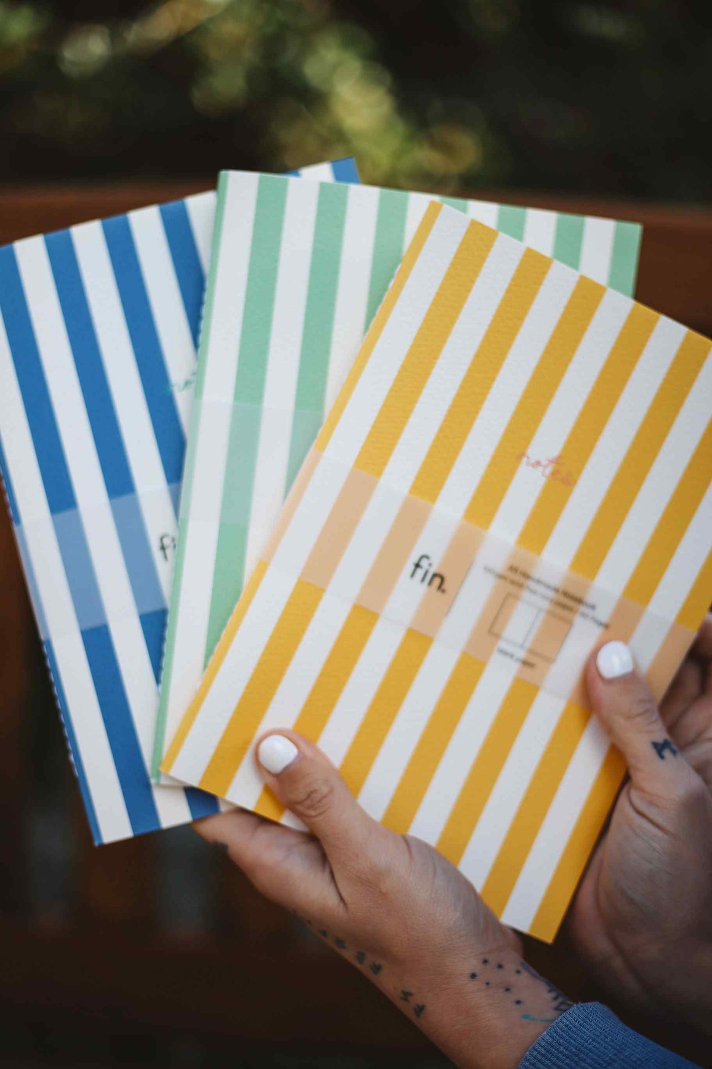Three striped notebook covers held in hands against a blurred outdoor background. The notebooks have vertical stripes in blue and white, mint green and white, and mustard yellow and white, with a small clear label on the front yellow notebook.
