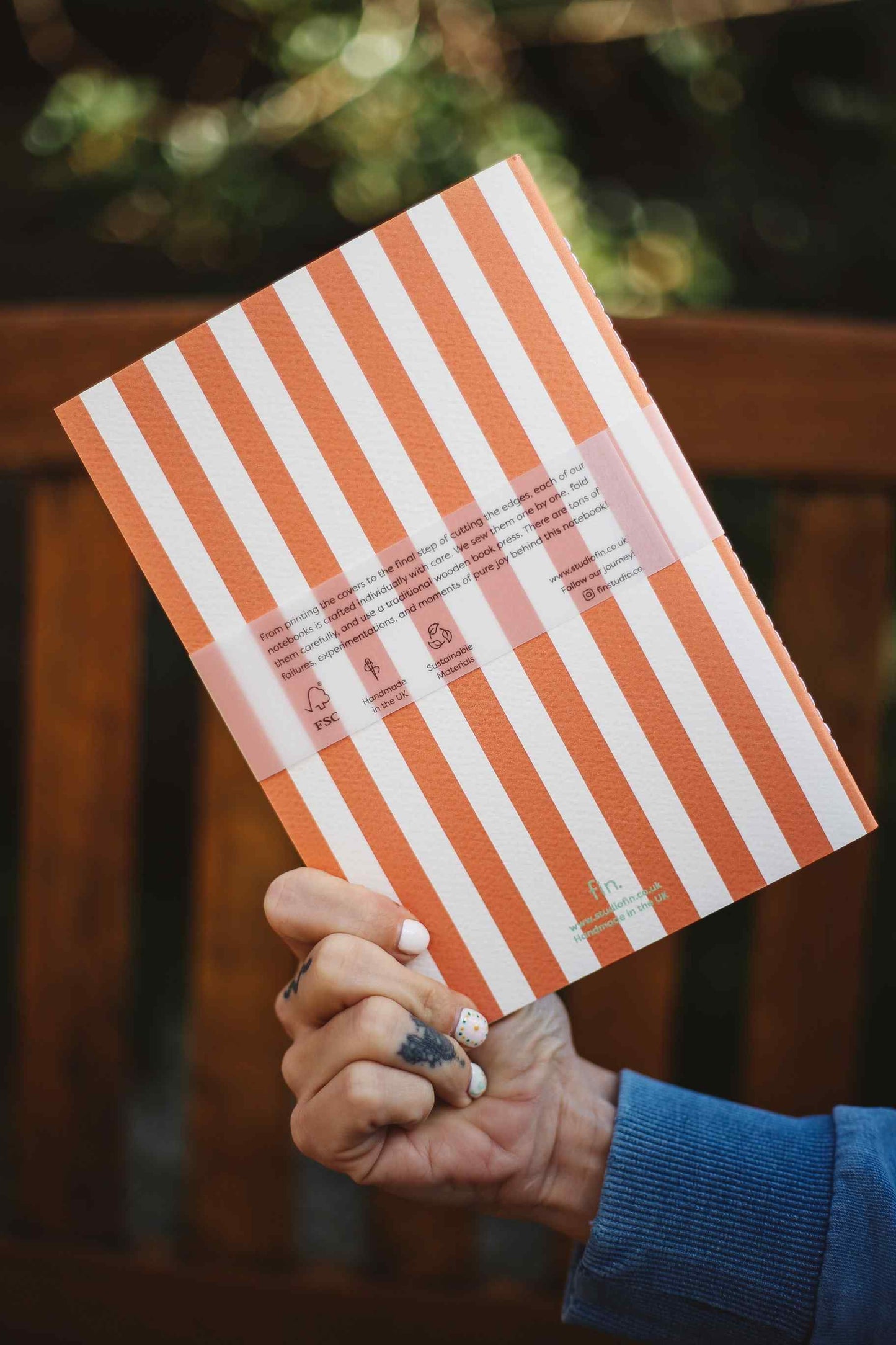 A hand holding a notebook with orange and white vertical stripes, featuring a translucent pink band across the middle with product details, set against a blurred outdoor background.