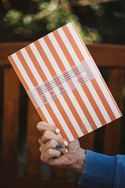 A hand holding a notebook with orange and white vertical stripes, featuring a translucent pink band across the middle with product details, set against a blurred outdoor background.