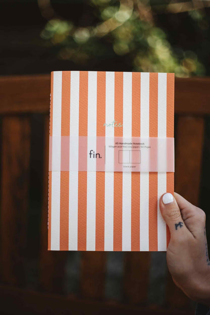 A hand with white nail polish holds a notebook with vertical orange and white stripes and a pink paper band labeled 'fin.' against a blurred dark wooden background.