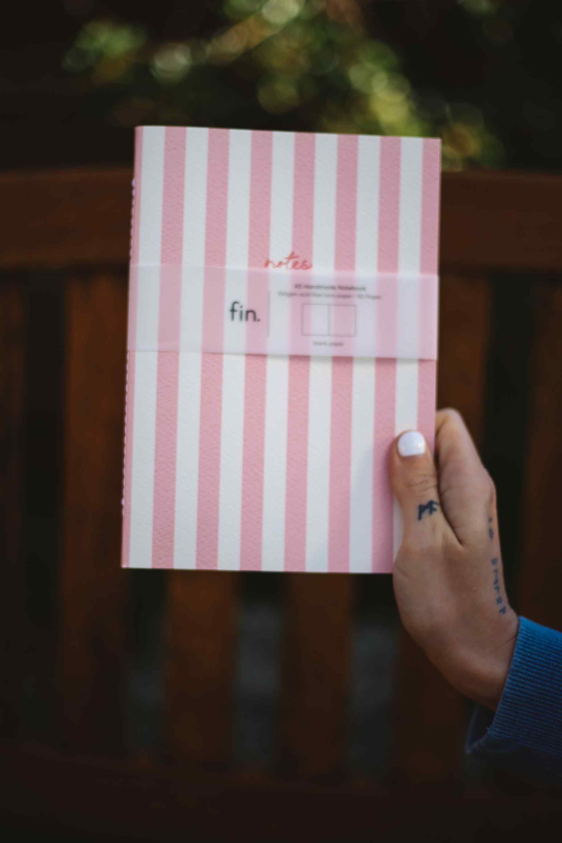 A hand with white nail polish holds a pink and white striped notebook with a paper band labeled 'notes fin.' against a dark wooden background.
