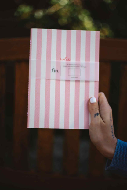 A hand with white nail polish holds a pink and white striped notebook with a paper band labeled 'notes fin.' against a dark wooden background.