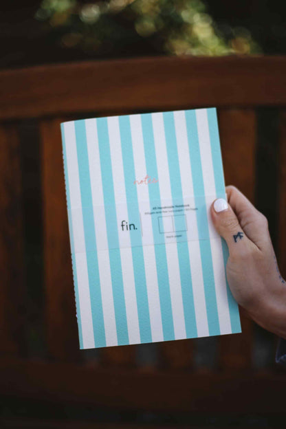 A hand with white nail polish holds a mint green and white striped envelope featuring the word 'fin.' in black text, set against a dark wooden background.